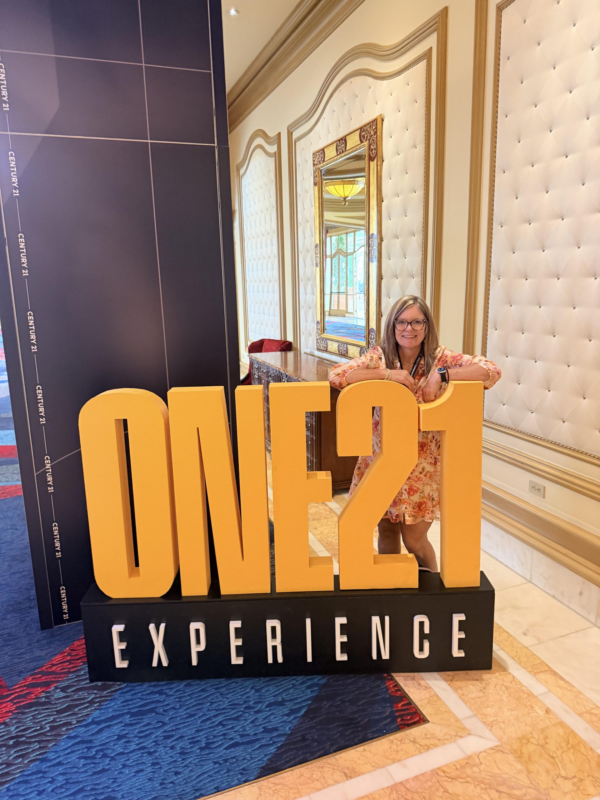 Kathy Paul standing behind the One21 Experience sign with a smile on her face